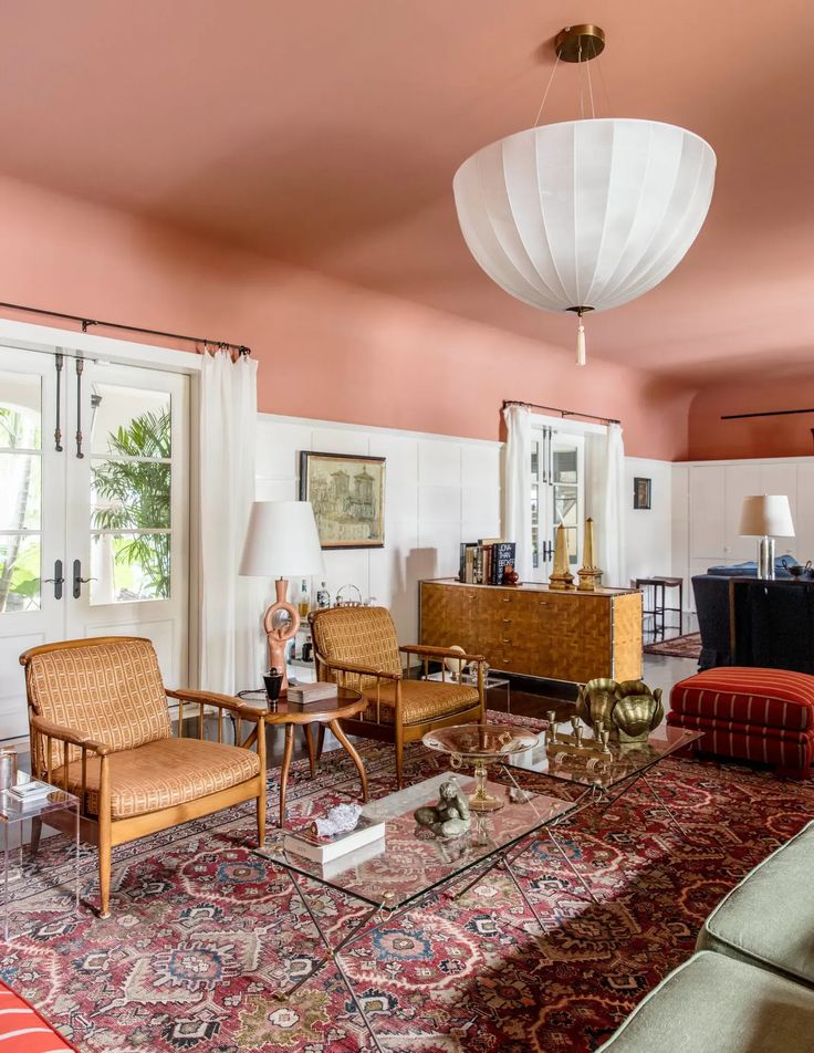 Living room with peach painted ceiling features a silk Josef Hoffmann chandelier from Woka, a pair of 1960s glass-and-brass Jacques Adnet coffee tables, midcentury Swedish armchairs and a late 19th-century Persian rug.
